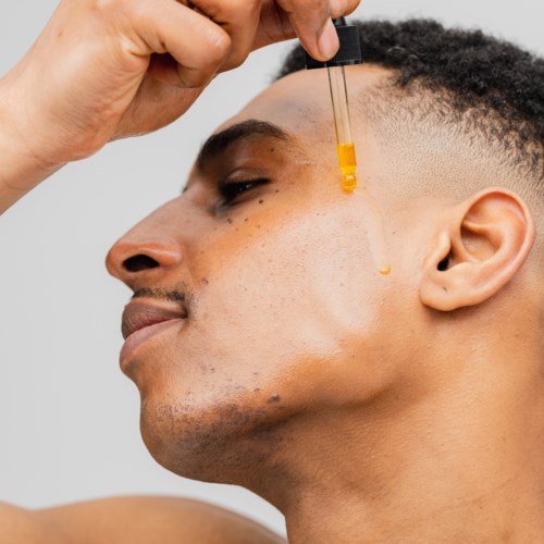 Ein Mann verwendet eine Pipette, um Öl auf sein Gesicht zu tropfen, mit kurzem Lockenkopf und ruhigem Ausdruck.