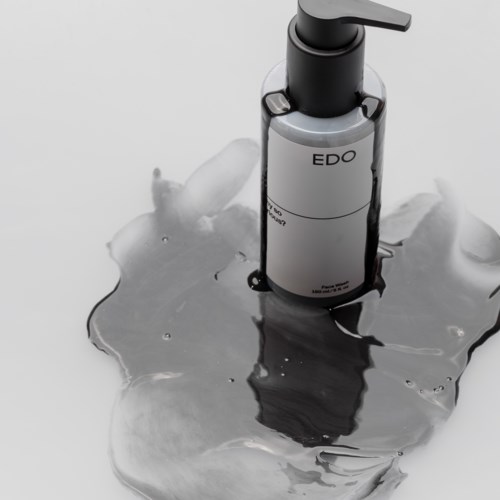 Eine graue Flasche mit Pumpaufsatz steht auf einer reflektierenden Oberfläche, mit verschütteter klarer Flüssigkeit.
