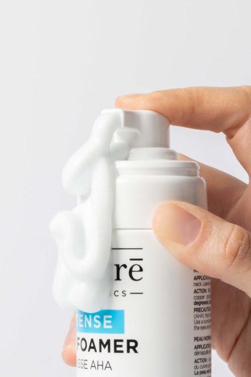 Eine Hand drückt eine Schaumpumpe auf eine weiße Hautpflegeflasche, mit Schaum, der über die Seiten läuft.
