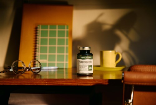 A desk with glasses, a green notebook, a supplement bottle, a yellow mug, and a brown bag in a cozy room.