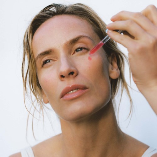 A woman uses a dropper to apply bright pink liquid on her face.