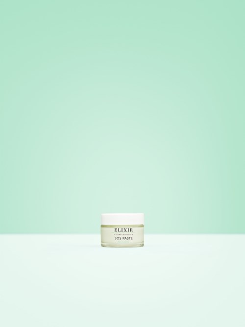 Ein kleiner weißer Behälter mit der Aufschrift 'Elixir SOS Paste' steht auf einer hellen Oberfläche vor einem mintgrünen Hintergrund.