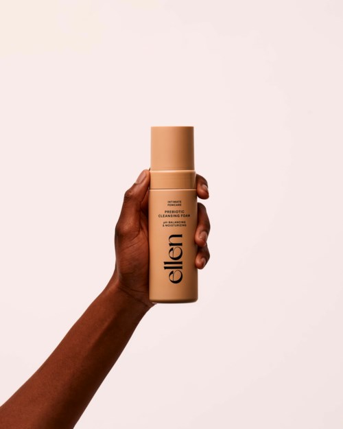 En hand håller en beige flaska med etiketten 'ellen' och enkel design mot en enkel bakgrund.