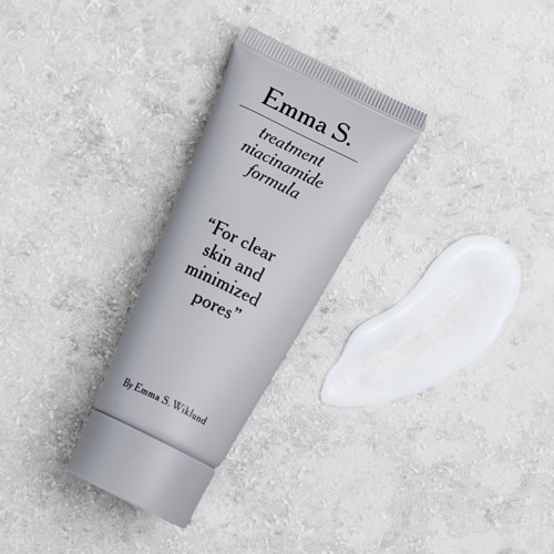 A white tube labeled Emma S., with cream beside it, on a light textured surface. The tube promotes pore-minimizing skin treatment.