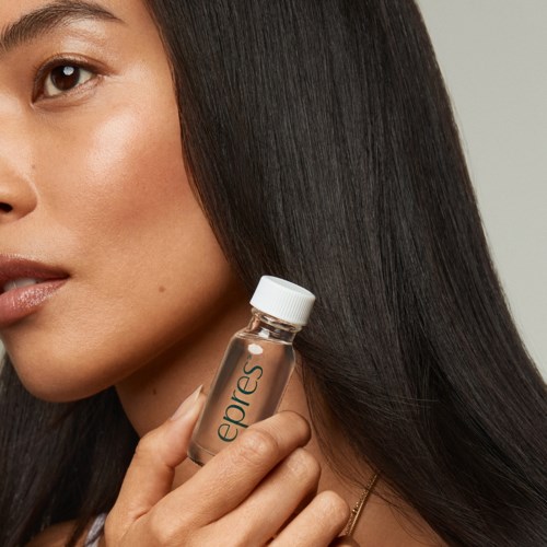 A woman with long, shiny black hair holding a small clear bottle close to her face.