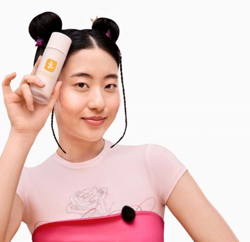 A woman with playful hair buns holds a skincare bottle next to her face, smiling against a white background.