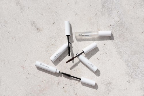 Vier witte mascara-flacons liggen verspreid op een lichte, gestructureerde ondergrond, met open en borstels zichtbaar.