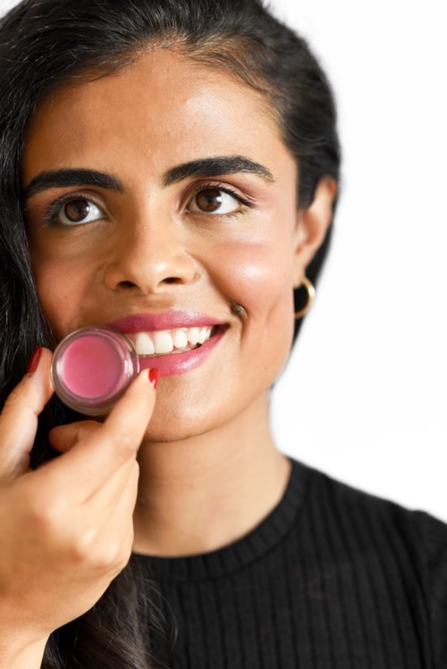 Een vrouw met donker haar die roze lipstick aanbrengt, glimlacht, draagt een zwarte top, op een witte achtergrond.