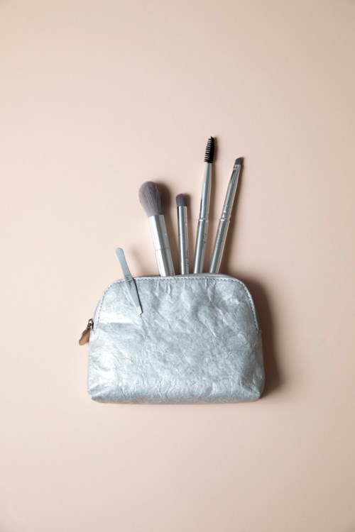 Een zilveren make-up tasje met vier make-up kwasten erin, tegen een eenvoudige lichte achtergrond.