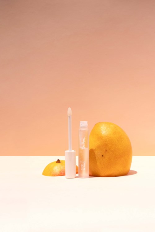 En persika, citron och läppbalsam med ett stift och en tub är placerade på en vit yta med en persikofärgad bakgrund.