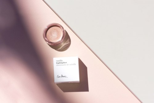 Een klein glazen potje en een doos met label vanille-highlighter liggen op een oppervlak met licht en schaduw.