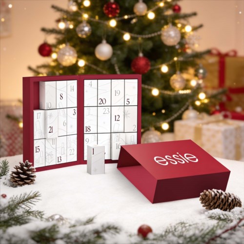 Rød adventskalender med 24 dager og 'essence'-logo, omgitt av kongler og julepynt på en snøhvit bord.