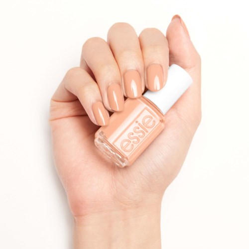 En hand med naglar målade i ljus persika, håller en persikofärgad flaska nagellack märkt 'essie'.
