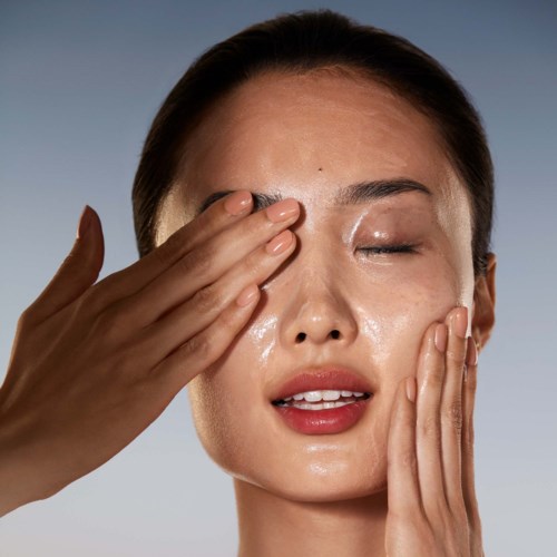A woman with shiny skin gently presses her face with both hands, closing her eyes, in a skincare routine or beauty context.