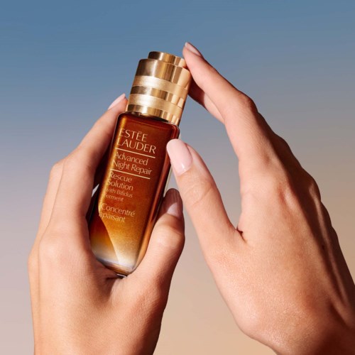 Een hand houdt een klein bruine fles met gouden dop, met label 'Estee Lauder Advanced Night Repair.'