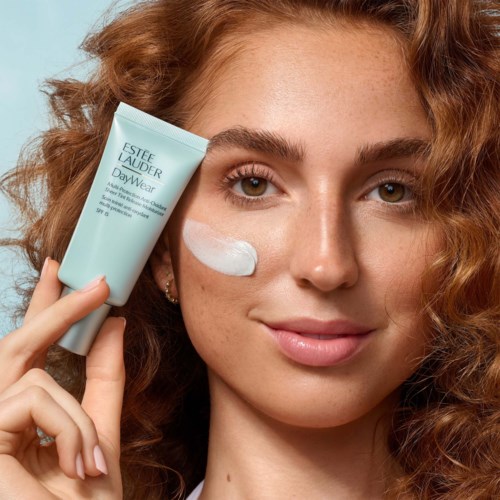 Een vrouw met gekruld rood haar smeert crème op haar wang met een tube die Estée Lauder aangeeft.