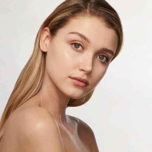 Een close-up van een jonge vrouw met lang blond haar en een heldere huid, die zacht naar de camera kijkt.