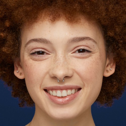 Leende ung kvinna med lockigt rött hår, fräknar och septum piercing, tittar mot kameran.
