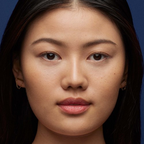 Een jonge vrouw met lang donker haar, een schone huid en minimale make-up, kijkt recht in de camera.