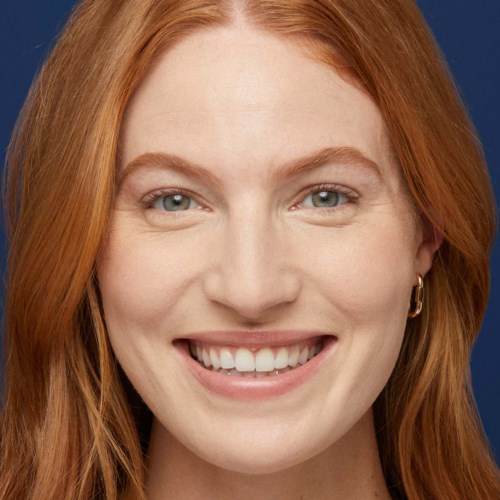 En ung kvinna med rött hår ler, bär örhängen, mot en mörkblå bakgrund.