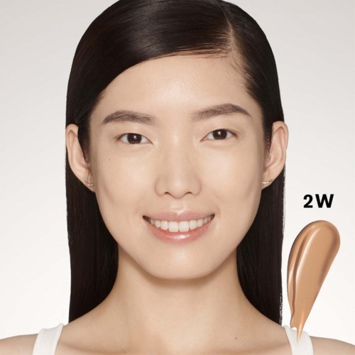 Een jonge vrouw met steil zwart haar en een vriendelijke glimlach, houdt een monster van beige foundation. De achtergrond is eenvoudig.