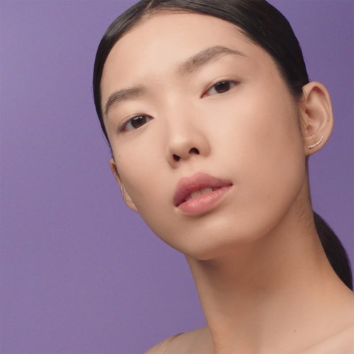 A woman with smooth skin and dark hair pulled back, looking calmly at the camera against a purple background.