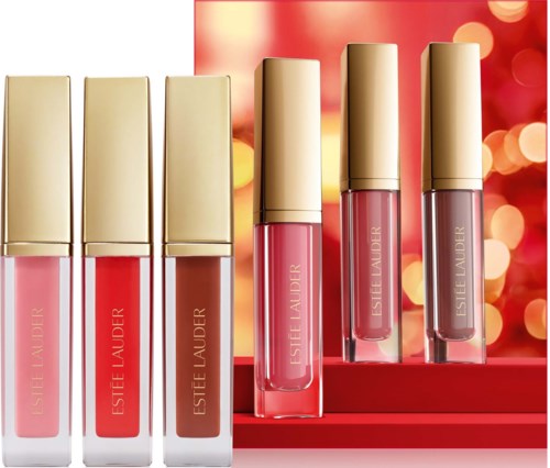 Six lip glosses in pink and nude shades arranged on a display with a festive red background and blurred lights.