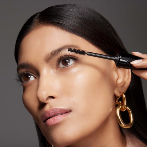 Vrouw brengt mascara aan op haar rechteroog, met recht zwart haar en grote gouden oorbellen.