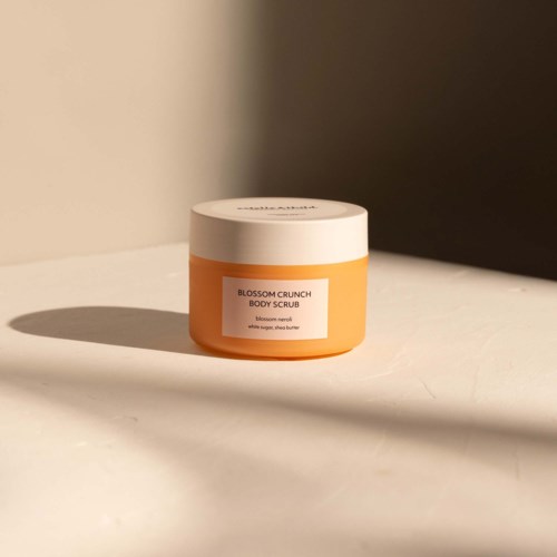 Een potje Blossom Crunch Body Scrub met oranje kleur, wit deksel en label. Het staat op een witte oppervlakte met schaduw.