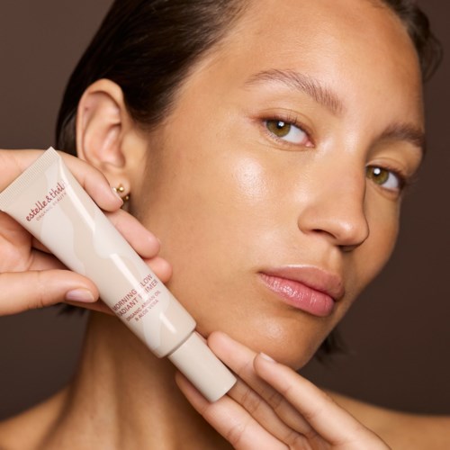 A woman with clear skin holds a tube of skincare product close to her face, looking at the camera.