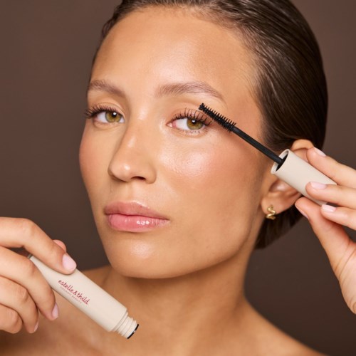 Woman applying mascara to her eyelashes, holding a white tube and using a mascara wand.