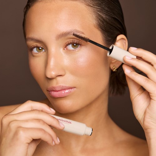 A woman applying mascara to her eyelash, holding the product with her right hand and looking at the camera.