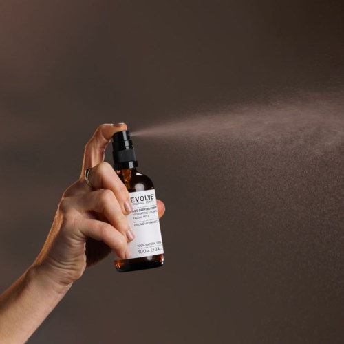 Eine Hand sprüht Flüssigkeit aus einer kleinen braunen Flasche mit Pumpspender auf die Haut. Der Hintergrund ist schlicht und dunkel.