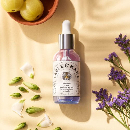 En rosa och lila glasflaska med Fable & Mane Amla lugnande serum med vit dropplock, omgiven av blommor.