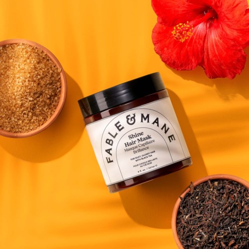 Fable & Mane Glow hårinpackning i burk med två små skålar med ingredienser, röd blomma nära, på ljusgul bakgrund.
