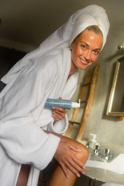 Eine Frau im Handtuch und mit Handtuch auf dem Kopf, lächelnd, hält Hautpflegeprodukte im Badezimmer.