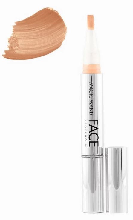 En concealerstift med ljus färg, med swatchad foundation i närheten och ett genomskinligt lock som reflekterar ljus.