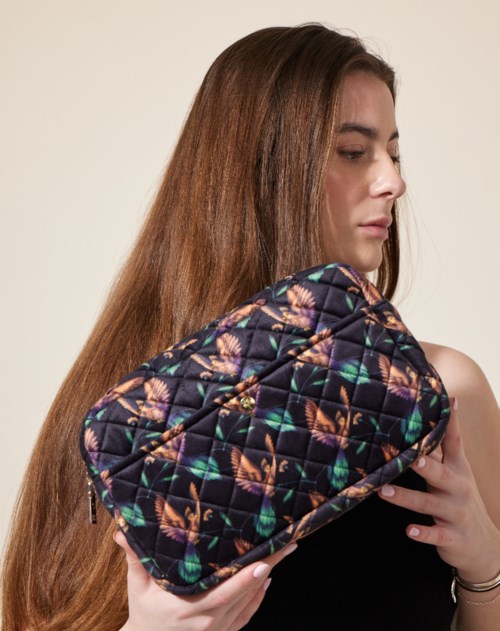 A woman with long brown hair holds a colorful, quilted bag with a peacock feather pattern against a plain background.