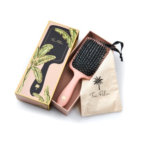 A pink hairbrush with black bristles, a box, and a cloth bag, all with palm tree designs for hair care.