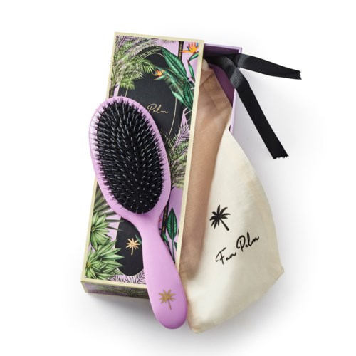 A purple hairbrush with a black bristle head rests on a tropical-themed box and a beige cloth bag with black ribbon and logo.