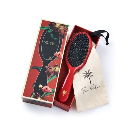 A red hairbrush with black bristles and a hanging loop, in a floral box and cloth pouch with palm tree design.