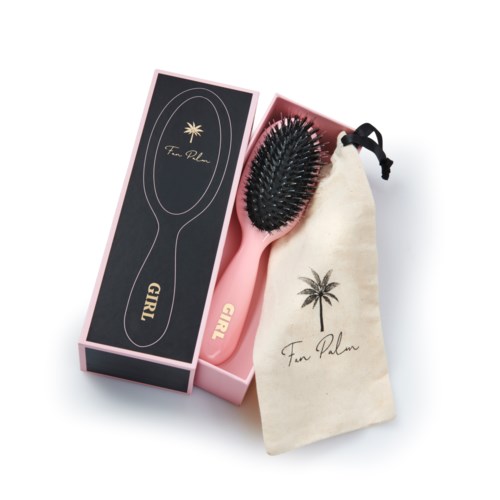 A pink hairbrush with black bristles and a black handle, in a pink box, accompanied by a cream bag with a palm tree logo.