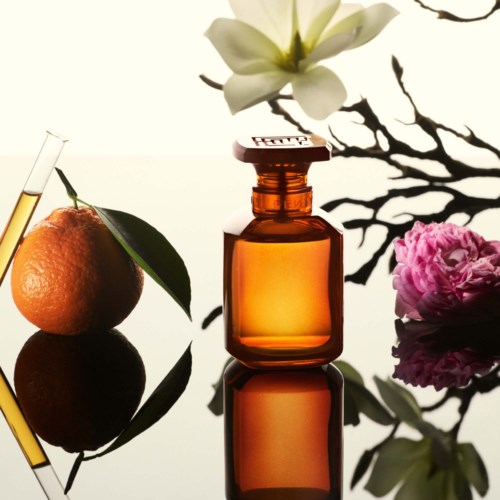A small amber bottle surrounded by a mandarin, a pink flower, and a white flower, all on a reflective surface.