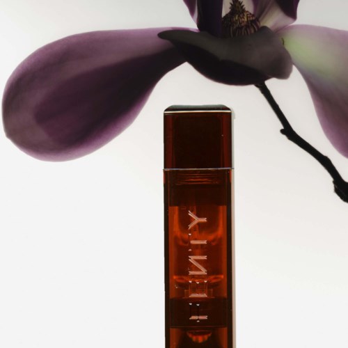 A brown glass bottle with the word 'LIT' written vertically, placed in front of a large, soft pink and purple flower.