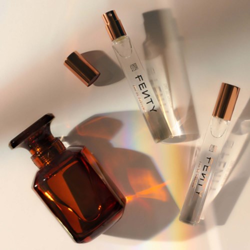 Two clear perfume vials and a brown bottle with a round stopper are on a white surface. Light creates rainbow reflections.