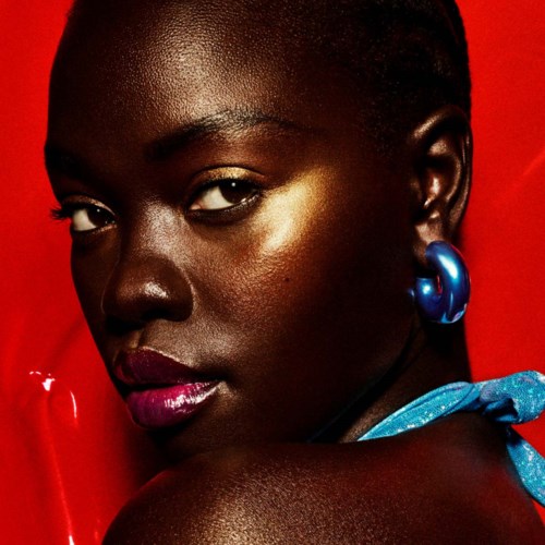 A woman with dark skin and painted lips, wearing blue earrings and a blue scarf, against a red background.