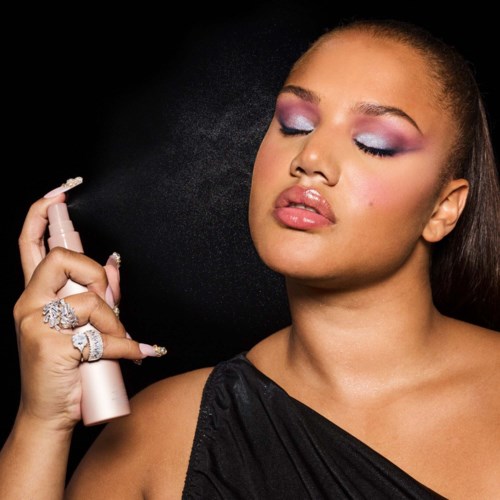 Woman with eye makeup, holding a spray bottle, wearing rings and a black outfit.
