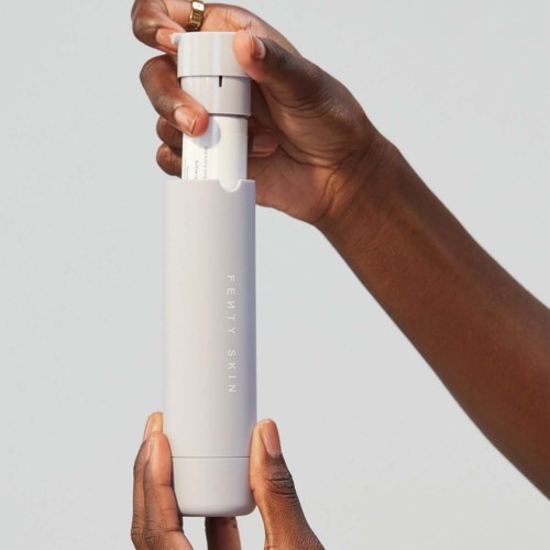 A hand holds a white cylindrical bottle labeled 'Fenty Skin' against a plain background.