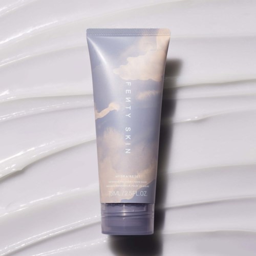 En tub med Fenty Skin-produkt med himmel- och molndesign på förpackningen. Den är placerad på en vit texturerad bakgrund.