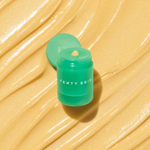 A green jar of Fenty Skin cream on shiny golden surface with open lid.